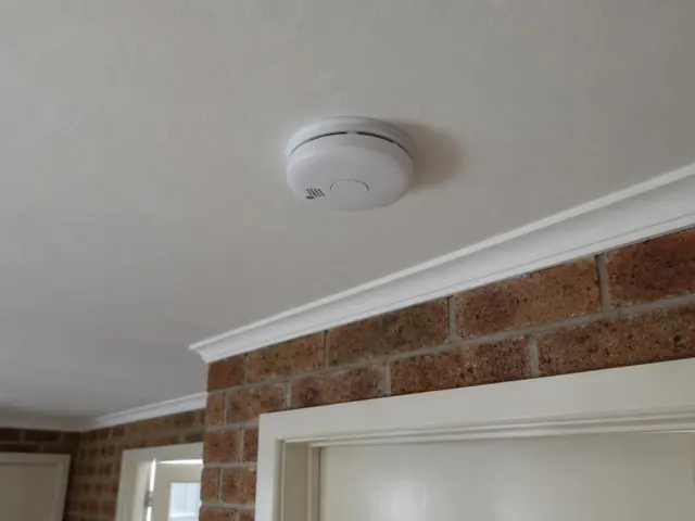 Ceiling-mounted smoke alarm above a brick interior wall and doorway in a home in Chittaway Bay